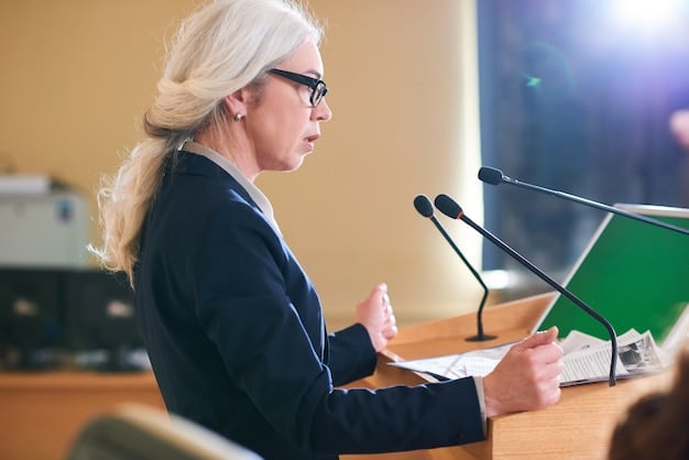 A close-up photo of Janet Yellen speaking at a podium during a Federal Reserve meeting, with charts and graphs projecting economic forecasts in the background.