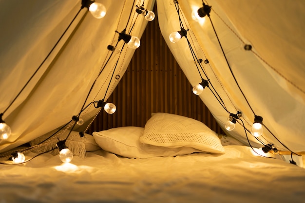 A close-up of the interior of a glamping tent, showing a comfortable bed with plush pillows, a rustic wooden nightstand, and a lantern providing soft lighting.
